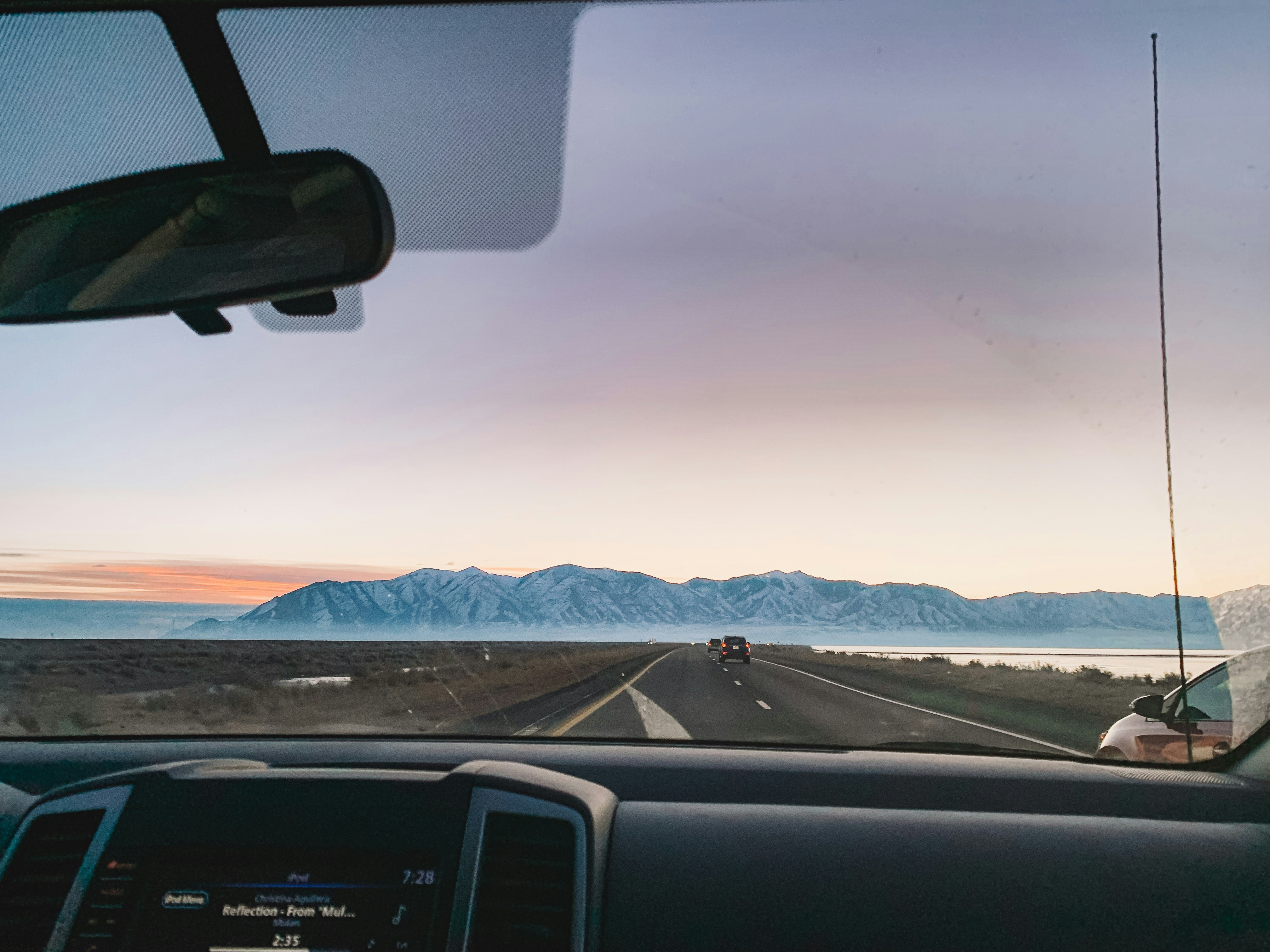 Dashboard view of a highway stretching toward snowy mountains at sunrise